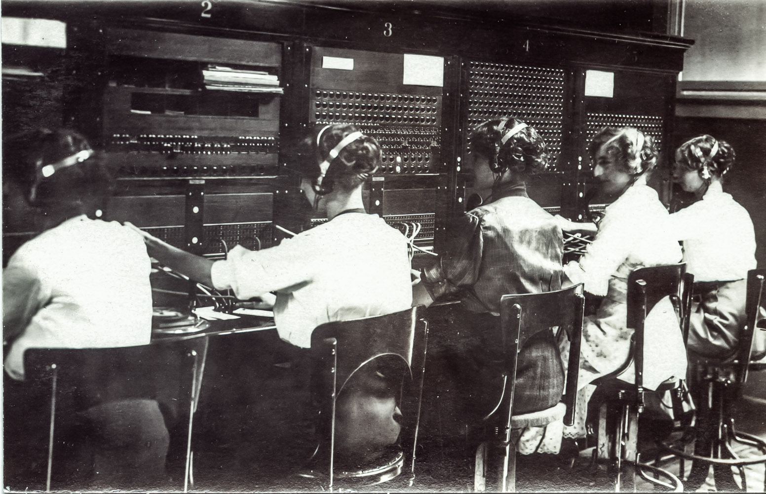 Early telephone operators wearing headsets at a switchboard, in black and white